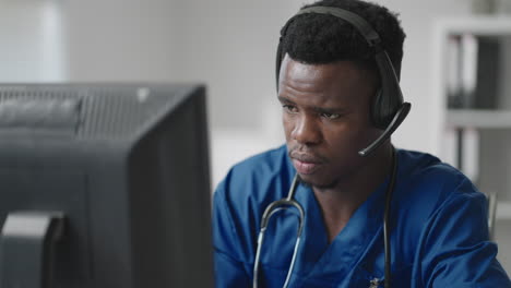 A-black-man-sits-at-a-computer-in-a-doctor's-uniform-and-writes-a-patient's-card-while-taking-calls-with-headphones.-Ambulance-Hotline-receive-calls-and-distribute-ambulances